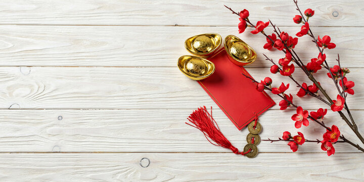 Chinese New Year background with red envelope, golden sycee ingots, coins tassel, and blooming red plum blossom branch on white wooden table.