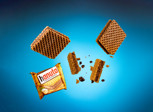 Creative studio composition of Ferrero Hanuta hazelnut wafers appearing suspended against a turquoise blue background, with a branded wrapper, broken wafer pieces, exposed cocoa and hazelnut filling, 