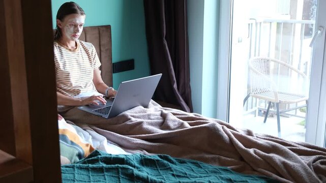 middle adult woman sitting in bed in facial clay mask using laptop for work or entertainment, morning routines, working from home