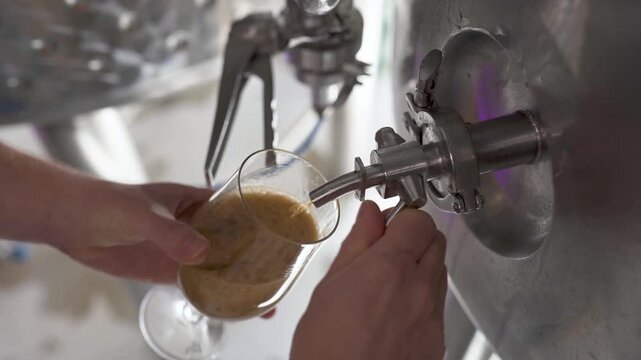 Fresh beer poured from stainless steel brewery tank into a glass, showing small batch brewing equipment and production process in an industrial brewery
