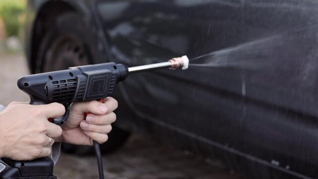 Close-up of a person using a pressure washer to clean a dirty car outdoors, removing dirt and grime with a powerful water jet.