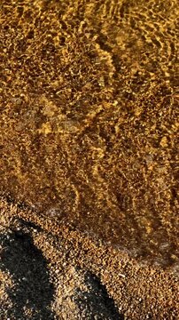 Gentle waves ripple along sunlit shoreline covered in golden pebbles. Moving seawater spreads across beach, forming brief white foam and tranquil pattern of amber reflections along waterline.