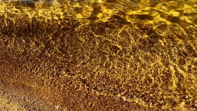 Sunlit seawater flows across pebbled shore, forming delicate ripples and thin lines of foam. Golden reflections shimmer over dark beach as gentle waves move back and forth, washing over wet stones.