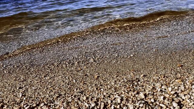 Gentle surf rolls across pebble beach, washing small stones before drawing back toward water. Rippling waves create peaceful shoreline pattern with clear shallows and scattered foam.