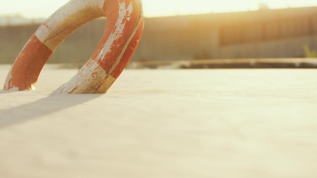 A lifebuoy with orange and white colors is placed on a wooden dock. The sun is setting in the background, casting warm light over the scene. The area is quiet and empty.