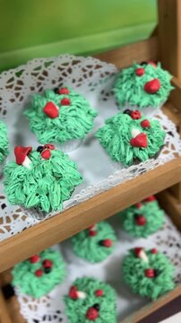 Green frosted cupcakes decorated with red berries and ladybugs. Displayed on wooden tiered tray with lace doily. Perfect for holiday baking or festive dessert scenes.
