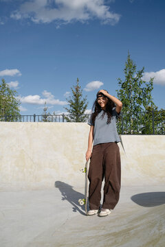 Skateboarder stands in a skate park under a blue sky with clouds