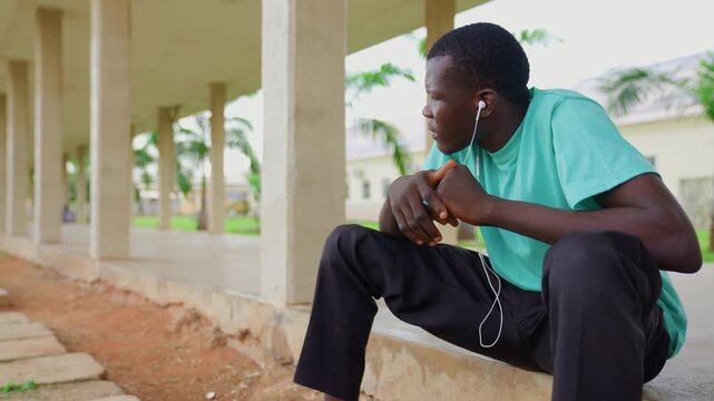 Teenage boy listens to audio through white earbuds while seated outdoors. Boy wears turquoise t shirt and black pants hands clasped in front of him under his chin