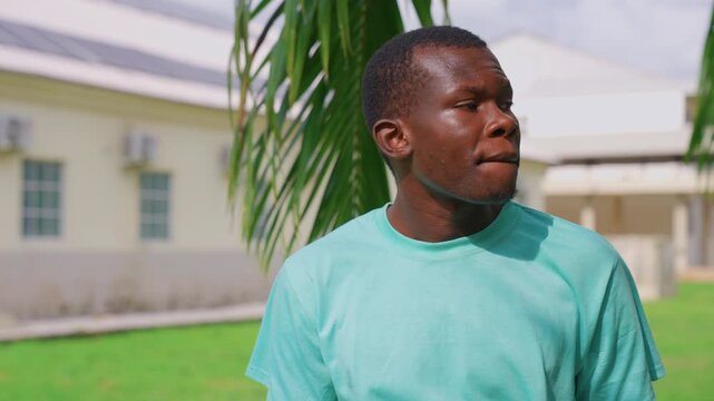 Young adult male wearing light blue shirt is positioned outside framed by green foliage from nearby plants. Behind him stands building with multiple windows and air conditioning units