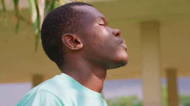 Close up side view shows back and right shoulder of young adult male with short curly hair wearing light blue t shirt. His head is tilted slightly upward at angle while his eyes are closed
