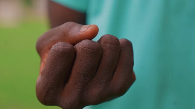 Close up view shows human hand starting in open position then gradually closing into fist. background is blurred green and subject wears teal garment emphasizing texture details