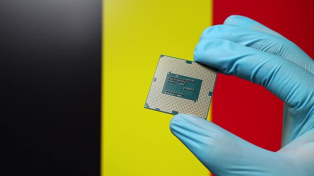 Close up view of hand in blue glove holding CPU microchip. Belgian flag in blurred background. Concept of semiconductor shortage and artificial intelligence development in Belgium.