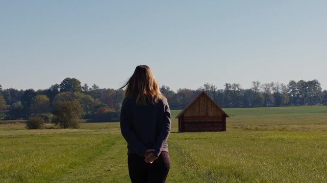 Woman walking through a scenic green meadow towards a traditional historic wooden hay barn in Trebon, Czech Republic