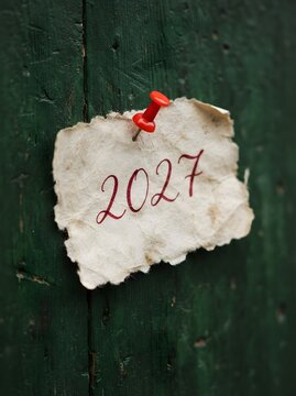 A small piece of white torn paper with the year 2027 written in red ink attached with a red push pin to a dark black wooden wall background for a new year celebration concept