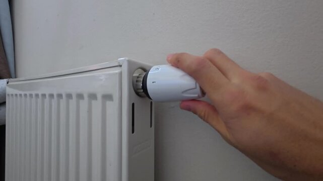 Turning thermostatic valve on radiator helps optimize indoor climate. Person regulates white heating controller by hand, managing residential temperature level for effective energy saving.