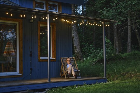 Middle-aged man sits on wooden chair on porch enjoying evening at cabin home. Surrounding trees create quiet space. String lights add warm glow as night approaches