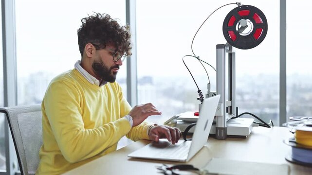 A focused man with curly hair and glasses works on a laptop next to a 3D printer with a spool of red filament