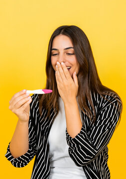 Woman reacts with surprise and joy to a positive pregnancy test against a yellow studio background. Image supports pregnancy announcement, fertility, family planning, motherhood and health campaigns.
