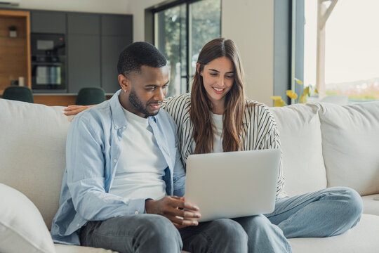 Couple uses a laptop together on a sofa at home, smiling as they browse online. Black man and Caucasian woman share technology in a bright living room with an open kitchen, enjoying connected life.