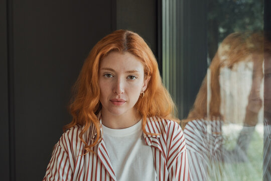 Redhead woman faces the camera beside a window, where her reflection doubles a direct gaze. Soft side light and dark interiors create a calm home portrait about identity, solitude, and self reflection