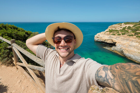 Relaxed man takes a selfie beside the rocky coast, adjusting his straw hat and smiling toward the camera. Blue ocean scenery evokes sunny vacation, coastal travel, tourism and summer relaxation.