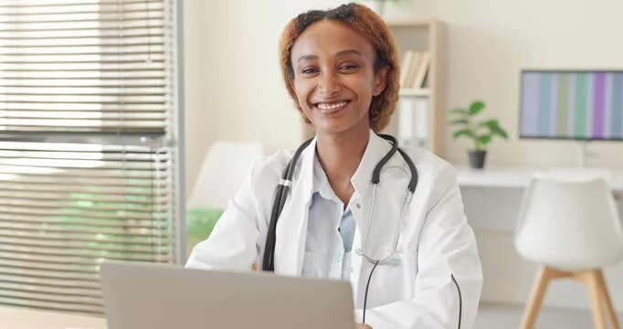 Smiling female doctor uses laptop in clinic. Friendly healthcare professional with stethoscope works at desk in bright medical office, supporting telehealth services. Trusted care concept.