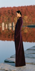 Fototapeta premium Side profile fashion image of woman in long plum coat standing on stone pier by autumn lake for editorial backdrop and lookbook