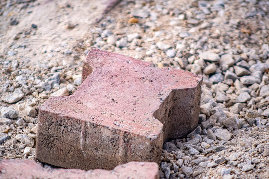 Broken paver block on gravel. A single chipped red concrete interlocking paver sits on crushed aggregate at a construction site. Suitable for construction, landscaping, and renovation projects.