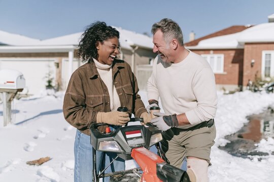 Snow blower shared by two laughing neighbours on wet asphalt driveways after a winter storm, for insurance and community media