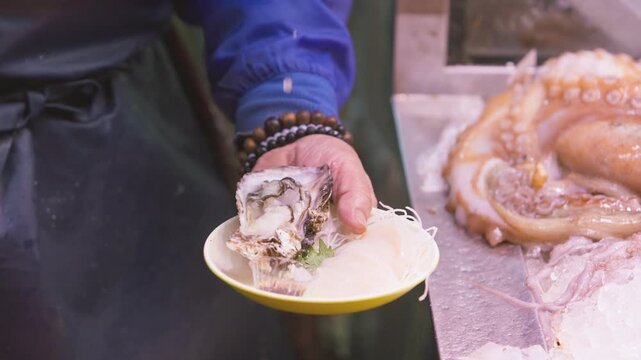 Captured in slow motion, a vendor squeezes lemon juice over an opened oyster on a yellow edged plate with fish slices and shredded garnish, then moves the plate from the counter toward a customer.