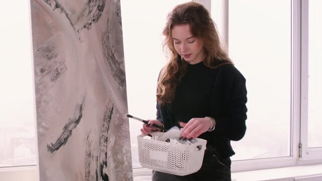 Acrylic painting. Creative workshop. Female professional master choosing color for abstract canvas near window in studio.