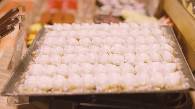 Captured in slow motion, pale steamed dumplings sit in rows on a tray above a heated food counter, with pleated wrappers and filling centers visible as steam drifts upward around the warm display.