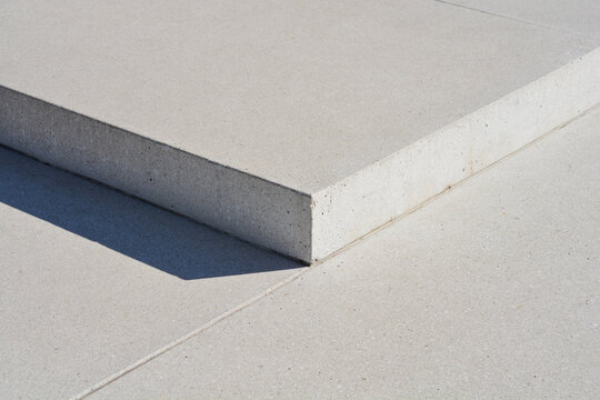 Architectural detail showing a minimalist light grey concrete step block casting a sharp shadow onto a smooth pavement surface with precise joint lines.