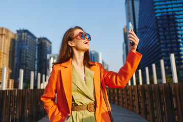 Fototapeta premium Stylish young woman in orange coat and sunglasses takes a selfie with smartphone against modern city skyscrapers. Concept of urban lifestyle, fashion and travel.