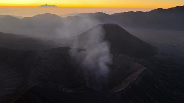 Volcano Mount Bromo emits smoke at sunrise. Rugged mountains frame the scene. Soft light bathes the crater