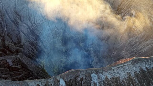 Drone aerial of Mount Bromo crater glowing with sunset light, steam and rugged volcanic textures.