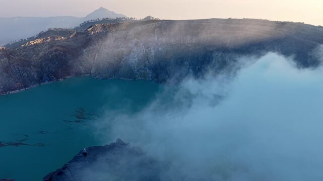 High altitude drone shot of Mount Ijen turquoise crater lake surrounded by steaming sulfur mist and rugged cliffs.