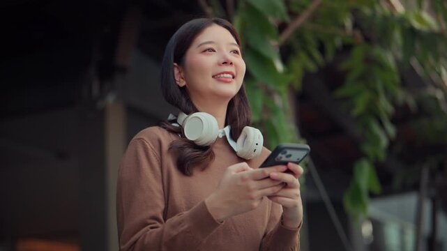 Happy young Asian woman smiling while using smartphone outdoors with wireless headphones around her neck, checking mobile messages