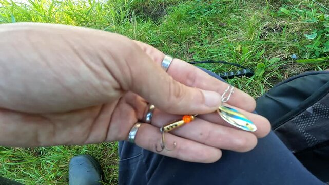 Close up POV of angler holding silver inline spinner fishing lure with treble hook. Fisherman preparing tackle on grassy river bank.