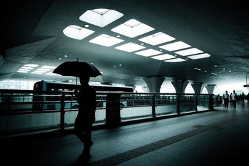 Fototapeta premium Silhouette of person with umbrella walks through dark urban tunnel. Modern architecture features bright skylights, geometric ceiling patterns. Public bus moves on street behind railing. Pedestrians
