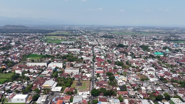 High altitude drone view showing Singosari town, rooftops, rice fields and distant mountains under bright daytime sky.