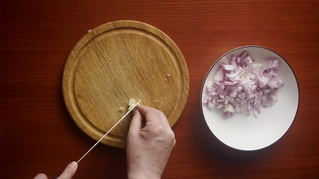 Horizontal Top-Down View of Hands Slicing Garlic Cloves next to Chopped Onions on Plate