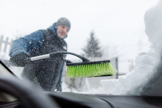 Man clearing fresh snow from windshield with brush, preparing vehicle for safe winter driving, view from inside vehicle