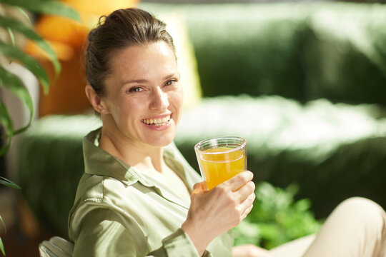 A bright, frontal portrait of a smiling woman in a green collared shirt holding a glass of orange juice, illustrating a vibrant and healthy start to the day.