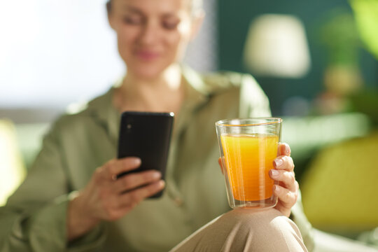 A middle-aged woman enjoys a moment of morning mindfulness, holding a fresh glass of orange juice and her smartphone in a warm, cozy home environment.
