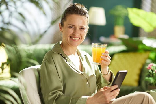 A smiling woman enjoys a glass of fresh orange juice while checking her phone in a sunlit, plant-filled living room, blending a healthy routine with digital connectivity.