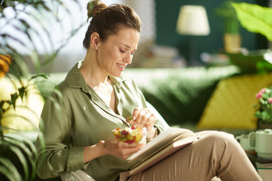 A smiling woman sits on a green sofa holding a bowl of fruit salad with a book on her lap, depicting a healthy lifestyle and clean eating during a relaxed morning at home.