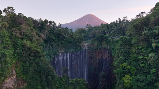 Majestic waterfall cascades through lush green canyon. Dense jungle wraps cliffs and misty gorge at Tumpak Sewu