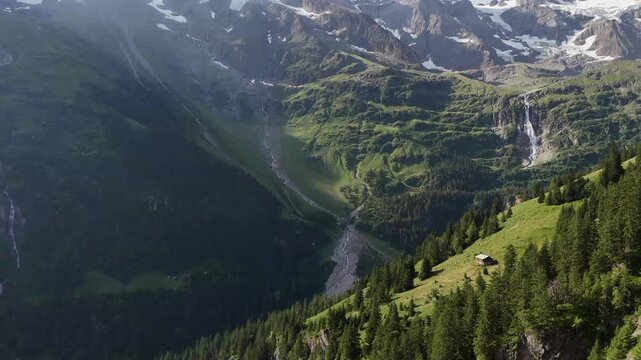 Drone view of a green alpine valley with forest, waterfall, rocky peaks, and lingering snow in the Swiss mountains.