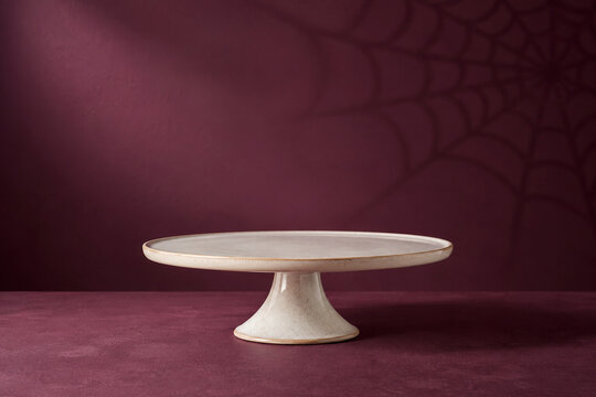 Empty white ceramic cake stand on a plum backdrop with a faint spiderweb shadow for Halloween product display